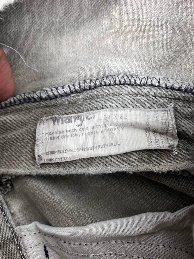 May include: Close-up of gray denim jeans with a white label. The label reads "Wrangler" and includes size information. The jeans have visible stitching and are made of 100% cotton.