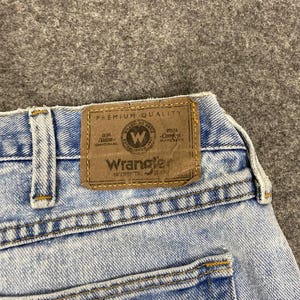 May include: Close-up of light blue denim jeans with a brown leather patch. The patch reads "Wrangler Authentic Jeans" with a circular logo. The jeans have yellow stitching and a classic five-pocket design.