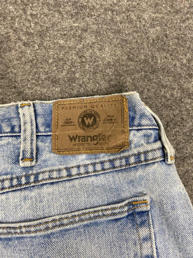 May include: Close-up of light blue denim jeans with a brown leather patch. The patch reads "Wrangler Authentic Jeans" with a circular logo. The jeans have yellow stitching and a classic five-pocket design.