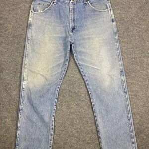 May include: Light blue denim jeans with a classic five-pocket design. The jeans show signs of wear and fading, with a slightly distressed hem. The jeans are laid flat on a gray surface.