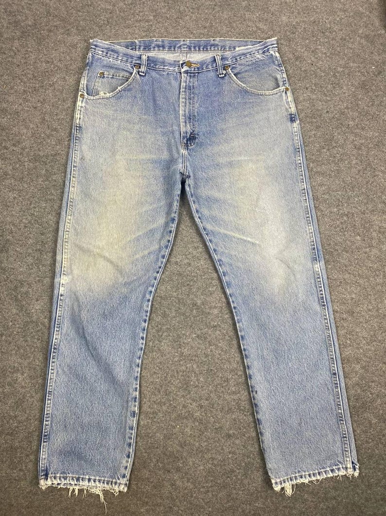 May include: Light blue denim jeans with a classic five-pocket design. The jeans show signs of wear and fading, with a slightly distressed hem. The jeans are laid flat on a gray surface.