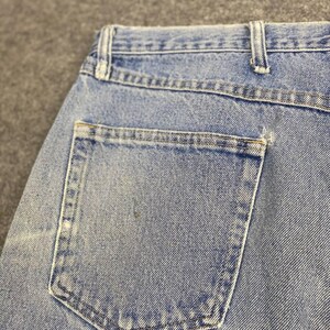 May include: Close-up of light blue denim jeans, showcasing the back pocket and waistband. The jeans have a classic design with visible stitching and a faded appearance, suggesting wear. The pocket is rectangular with reinforced stitching.