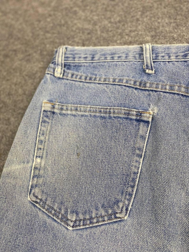 May include: Close-up of light blue denim jeans, showcasing the back pocket and waistband. The jeans have a classic design with visible stitching and a faded appearance, suggesting wear. The pocket is rectangular with reinforced stitching.