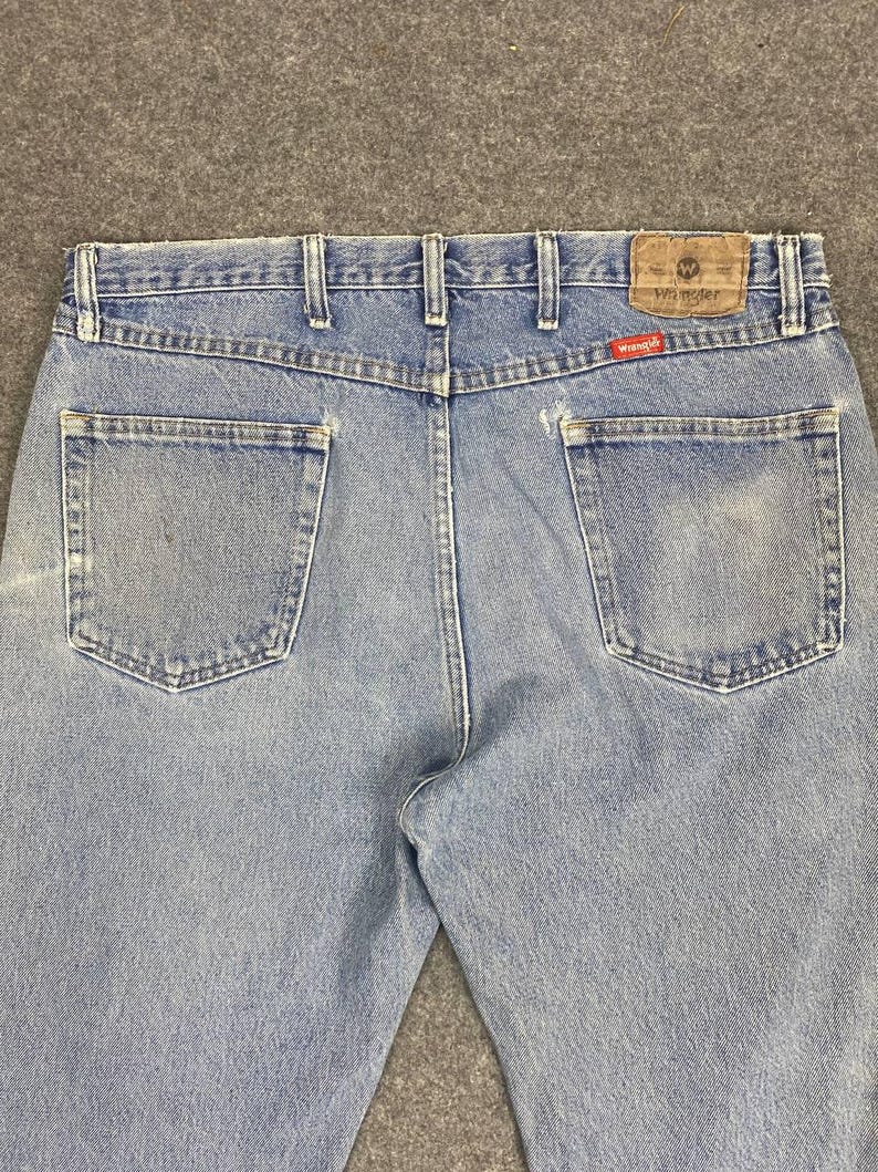 May include: Light blue denim jeans with two back pockets and a brown leather patch. The jeans feature the Wrangler logo on the patch and a red tag. The jeans have belt loops and a classic five-pocket design.