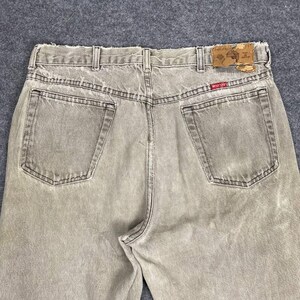 May include: Back view of faded tan denim jeans with two back pockets and a leather patch. The jeans have belt loops and a red and brown label. The denim has a worn, vintage look.