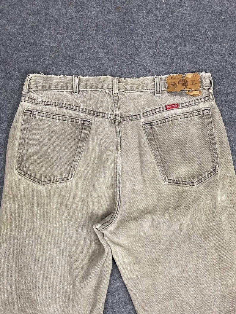 May include: Back view of faded tan denim jeans with two back pockets and a leather patch. The jeans have belt loops and a red and brown label. The denim has a worn, vintage look.