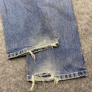 May include: Close-up of light blue denim jeans with distressed details. The jeans have a faded appearance with frayed edges at the hem, showing signs of wear. The fabric is textured, and the overall look is casual and vintage.