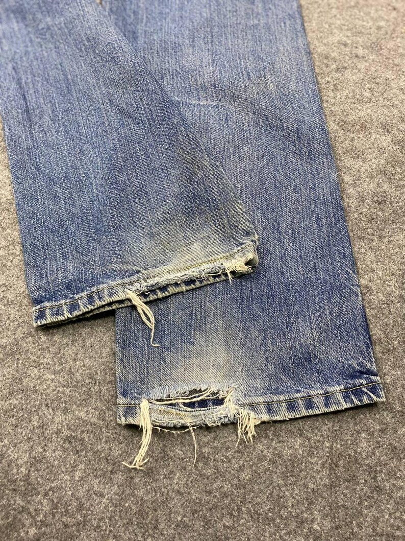 May include: Close-up of light blue denim jeans with distressed details. The jeans have a faded appearance with frayed edges at the hem, showing signs of wear. The fabric is textured, and the overall look is casual and vintage.