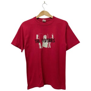 May include: Red t-shirt with a graphic featuring the band name "THE FLIPSIDES" in black letters. The graphic includes three silhouetted figures. The shirt is hanging on a wooden hanger.