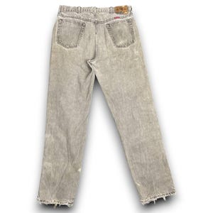 May include: Back view of light gray denim jeans with a faded, distressed appearance. The jeans have a classic five-pocket design, a button and zipper closure, and a leather patch on the waistband. The hems are frayed.