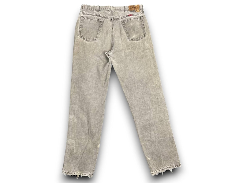 May include: Back view of light gray denim jeans with a faded, distressed appearance. The jeans have a classic five-pocket design, a button and zipper closure, and a leather patch on the waistband. The hems are frayed.