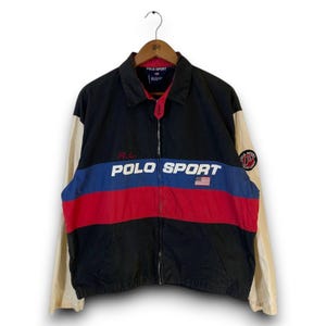 May include: A black, red, and blue color-blocked windbreaker jacket with cream-colored sleeves. The front features the text POLO SPORT in white, a small American flag graphic, and a red embroidered RL logo. A circular Polo Sport patch is on the sleeve.