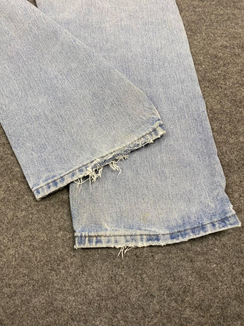 May include: Close-up of light blue denim jeans. The bottom hems are frayed and distressed, with visible threads. The jeans show signs of wear and tear, with slight discoloration.