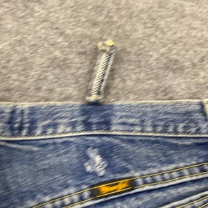 May include: Close-up of blue denim jeans with yellow stitching. The jeans have a button and a zipper. The Wrangler brand label is visible on the back pocket. The jeans are on a gray surface.