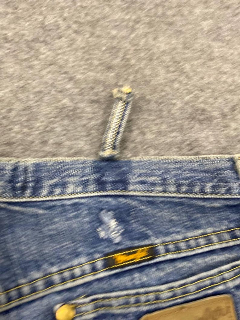 May include: Close-up of blue denim jeans with yellow stitching. The jeans have a button and a zipper. The Wrangler brand label is visible on the back pocket. The jeans are on a gray surface.