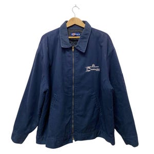 May include: Navy blue work jacket with a full zipper closure and a collared neckline. The jacket features a small white logo on the chest that reads "IC Construction". The jacket is hanging on a wooden hanger.