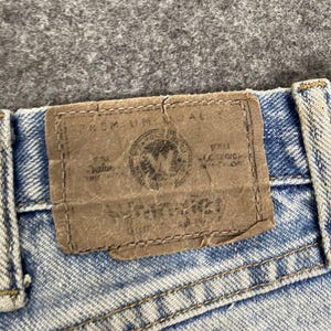 K&ouml;nnte beinhalten: Nahaufnahme einer hellblauen Jeans mit einem braunen Lederaufn&auml;her. Der Aufn&auml;her hat gepr&auml;gten Text und ein kreisf&ouml;rmiges Emblem. Die Jeans hat sichtbare N&auml;hte und ein verwaschenes Aussehen.