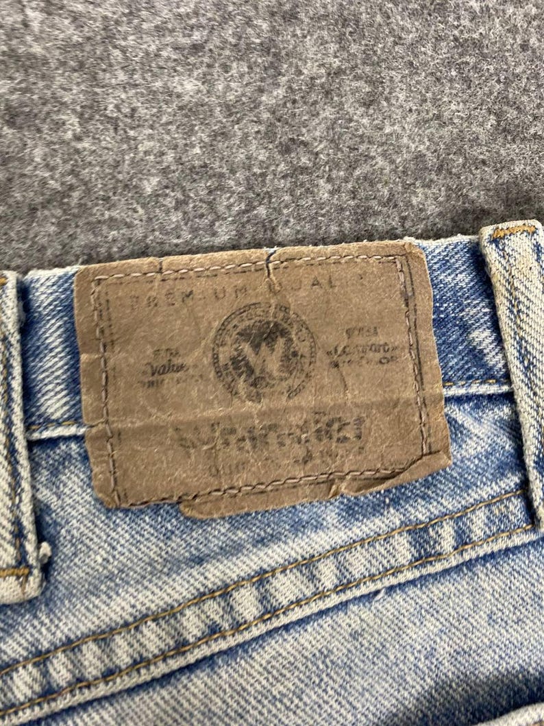 K&ouml;nnte beinhalten: Nahaufnahme einer hellblauen Jeans mit einem braunen Lederaufn&auml;her. Der Aufn&auml;her hat gepr&auml;gten Text und ein kreisf&ouml;rmiges Emblem. Die Jeans hat sichtbare N&auml;hte und ein verwaschenes Aussehen.