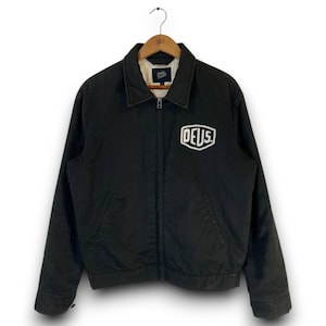 Könnte beinhalten: Schwarze Jacke mit Kragen und Reißverschluss. Auf der Vorderseite der Jacke befindet sich ein weißes "DEUS"-Logo. Sie hängt an einem Holzkleiderbügel.