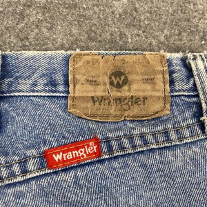 May include: Close-up of blue denim jeans with a brown leather patch and a red Wrangler tag. The leather patch has the Wrangler logo and text. The red tag is stitched on the back pocket.