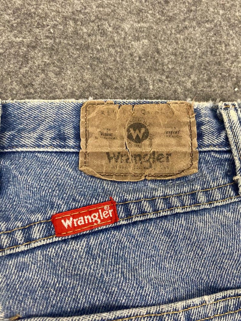 May include: Close-up of blue denim jeans with a brown leather patch and a red Wrangler tag. The leather patch has the Wrangler logo and text. The red tag is stitched on the back pocket.