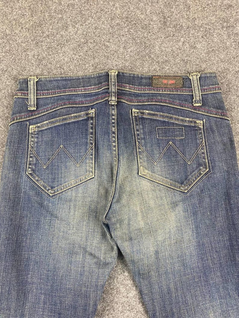 Puede incluir: Primer plano de la parte trasera de unos vaqueros azules. Los vaqueros tienen dos bolsillos traseros con un dise&ntilde;o de 'W' cosido. Las costuras son de color rosa en contraste. Una peque&ntilde;a etiqueta de cuero es visible en la cintura.