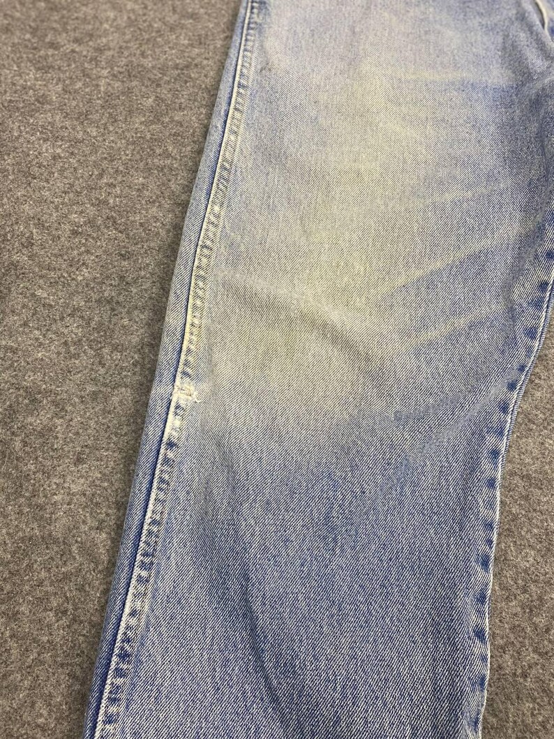 May include: Close-up of light blue denim jeans. The jeans show signs of wear with faded areas and visible stitching along the seams. The fabric has a slightly distressed appearance, suggesting a vintage or well-worn style.