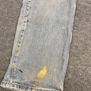 K&ouml;nnte beinhalten: Nahaufnahme einer hellblauen Jeans mit Gebrauchsspuren. Die Jeans hat mehrere Flecken und Markierungen, darunter ein gro&szlig;er gelber Fleck in der N&auml;he des Saums. Der Stoff weist Verblassungen und Abnutzungserscheinungen auf.