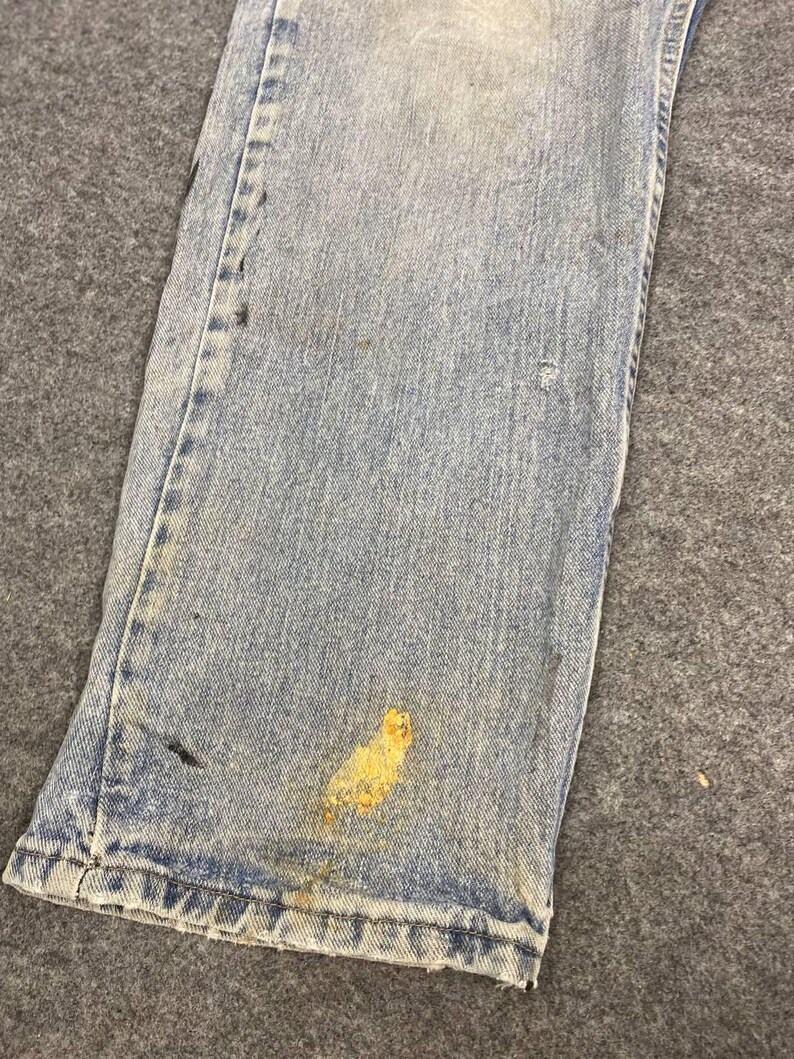 K&ouml;nnte beinhalten: Nahaufnahme einer hellblauen Jeans mit Gebrauchsspuren. Die Jeans hat mehrere Flecken und Markierungen, darunter ein gro&szlig;er gelber Fleck in der N&auml;he des Saums. Der Stoff weist Verblassungen und Abnutzungserscheinungen auf.