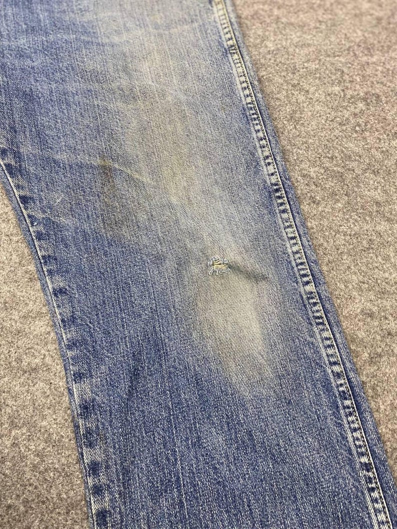 May include: Close-up of light blue denim jeans with a distressed appearance. The jeans show signs of wear, including a small tear and discoloration. The stitching is visible along the seams.