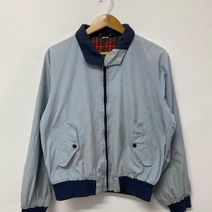 Könnte beinhalten: Hellblaue Jacke mit marineblauem Kragen, Bündchen und Saum. Die Jacke hat einen durchgehenden Reißverschluss, zwei Vordertaschen und ein kariertes Futter, das am Kragen sichtbar ist. Hängt an einem Holzbügel.