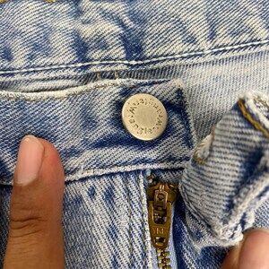 May include: Close-up of light blue denim jeans with a silver button and a brass zipper. The button is embossed with the word "Wrangler". The zipper is marked "YKK".