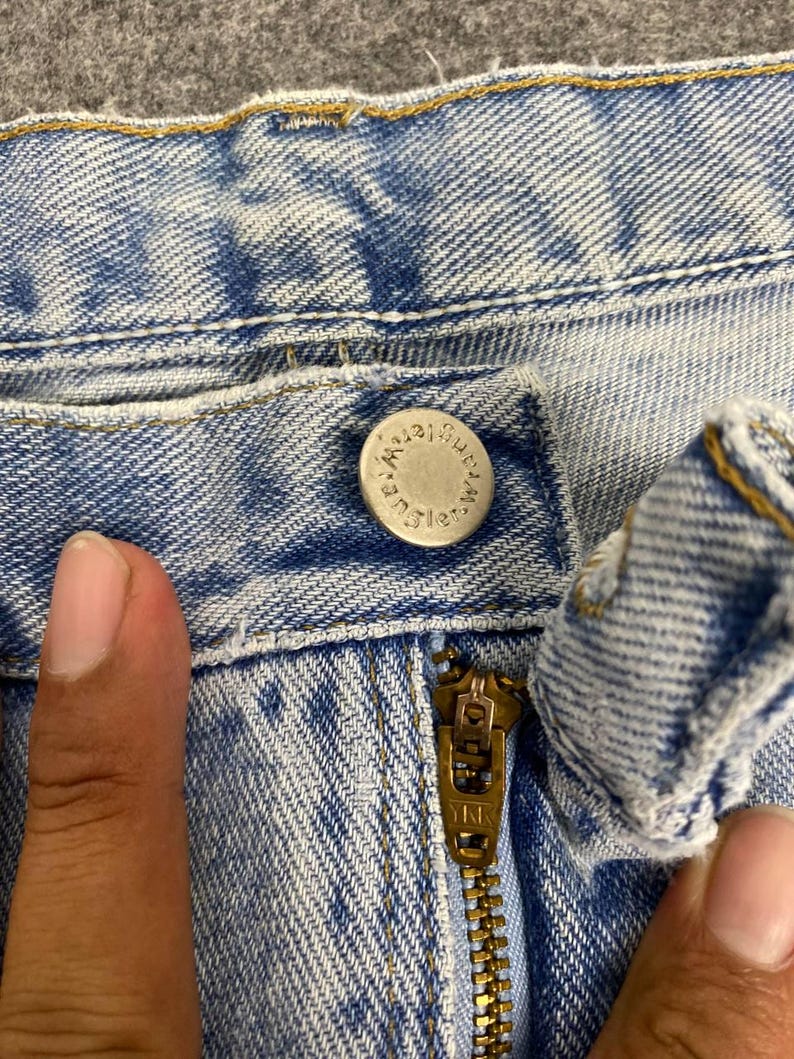 May include: Close-up of light blue denim jeans with a silver button and a brass zipper. The button is embossed with the word "Wrangler". The zipper is marked "YKK".