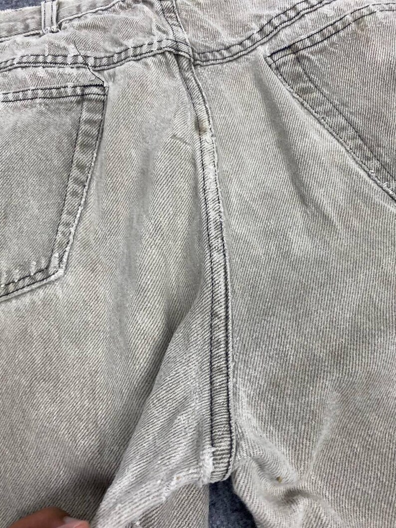 May include: Close-up of light gray denim jeans. The jeans have visible stitching in black thread along the seams and pockets. The fabric has a textured appearance with vertical lines.
