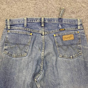 May include: Close-up of blue denim Wrangler jeans. The jeans feature classic five-pocket styling, a brown leather patch with the Wrangler logo, and signature stitching on the back pockets. The jeans have a button and zipper closure.