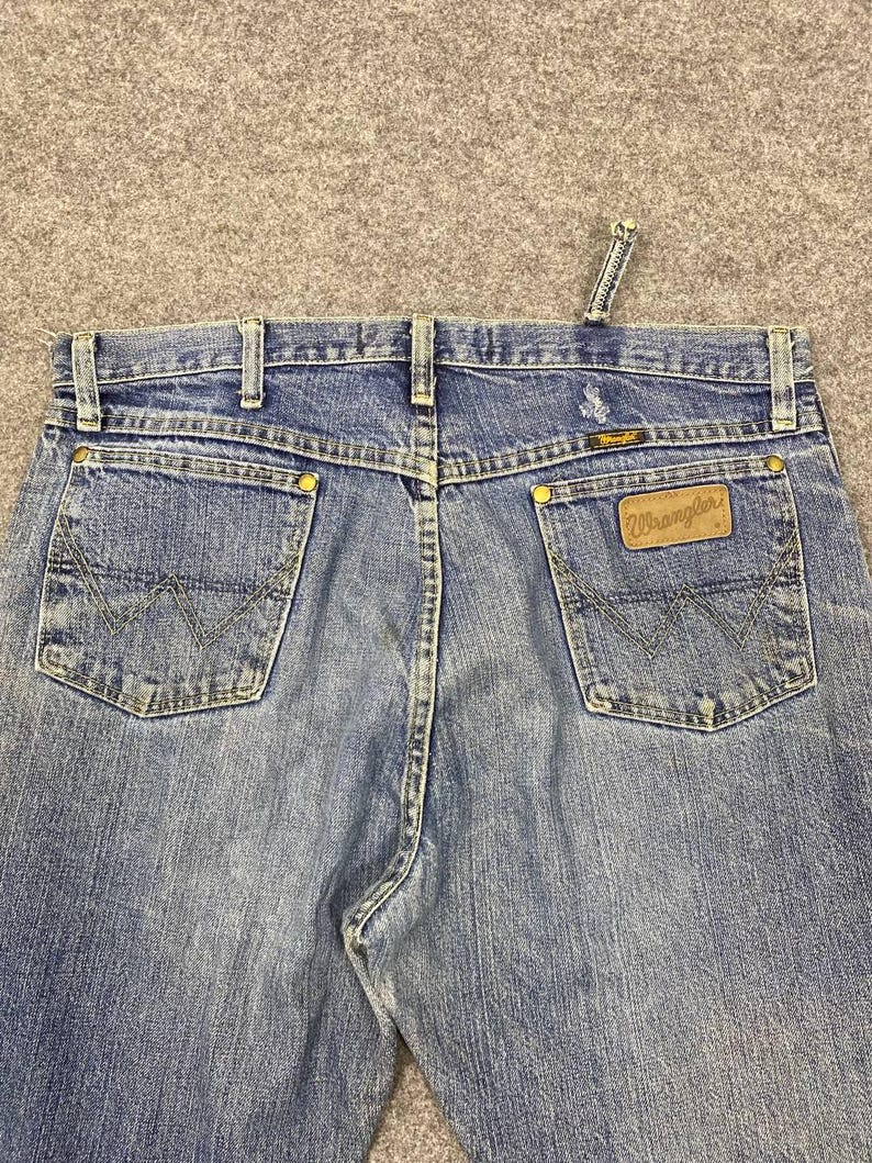 May include: Close-up of blue denim Wrangler jeans. The jeans feature classic five-pocket styling, a brown leather patch with the Wrangler logo, and signature stitching on the back pockets. The jeans have a button and zipper closure.