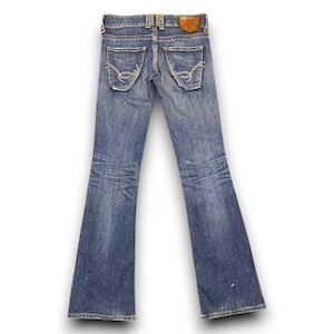 Puede incluir: Vaqueros de mezclilla azul descolorido con pernera acampanada. Los vaqueros tienen un diseño clásico de cinco bolsillos, con costuras decorativas en los bolsillos traseros. Un parche de cuero marrón es visible en la cintura. Los vaqueros se muestran sobre un fondo blanco.