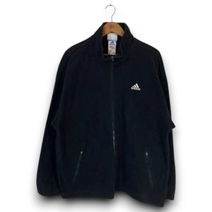 Peut inclure: Veste polaire Adidas noire à fermeture éclair intégrale avec col montant. Comporte un logo Adidas blanc sur la poitrine et deux poches avant. La veste est suspendue à un cintre en bois.