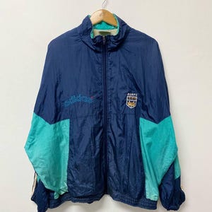 May include: Vintage Adidas track jacket in navy blue and teal. The jacket features a full zip, a stand-up collar, and the Adidas logo embroidered on the chest. The jacket also has the text "AURFC" and a crest on the chest.