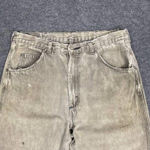 May include: Close-up of faded, light brown denim jeans. The jeans have a classic five-pocket design, a button and zipper closure, and belt loops. The fabric shows signs of wear and tear, with distressed details.