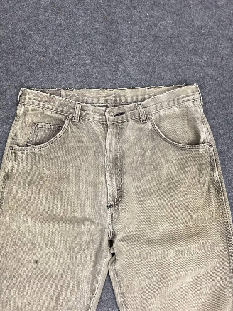 May include: Close-up of faded, light brown denim jeans. The jeans have a classic five-pocket design, a button and zipper closure, and belt loops. The fabric shows signs of wear and tear, with distressed details.