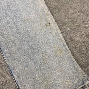 K&ouml;nnte beinhalten: Nahaufnahme eines Abschnitts einer hellblauen Jeans. Der Stoff weist Abnutzungserscheinungen mit mehreren braunen Flecken auf. Die Jeans ist gefaltet und zeigt den Saum und die Textur des Denim.
