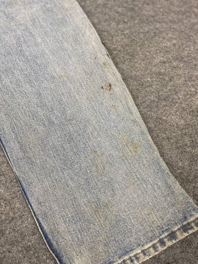 K&ouml;nnte beinhalten: Nahaufnahme eines Abschnitts einer hellblauen Jeans. Der Stoff weist Abnutzungserscheinungen mit mehreren braunen Flecken auf. Die Jeans ist gefaltet und zeigt den Saum und die Textur des Denim.