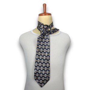 May include: A dark blue necktie featuring a repeating pattern of light tan and white oval shapes. The tie is tied in a knot around the neck of a white mannequin torso. The fabric has a smooth, slightly reflective finish with a pointed bottom end.
