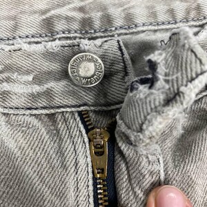May include: Close-up of the top portion of a pair of light gray denim jeans. The jeans feature a metal button with the "Wrangler" logo, a brass zipper, and visible stitching details. The fabric shows signs of wear.