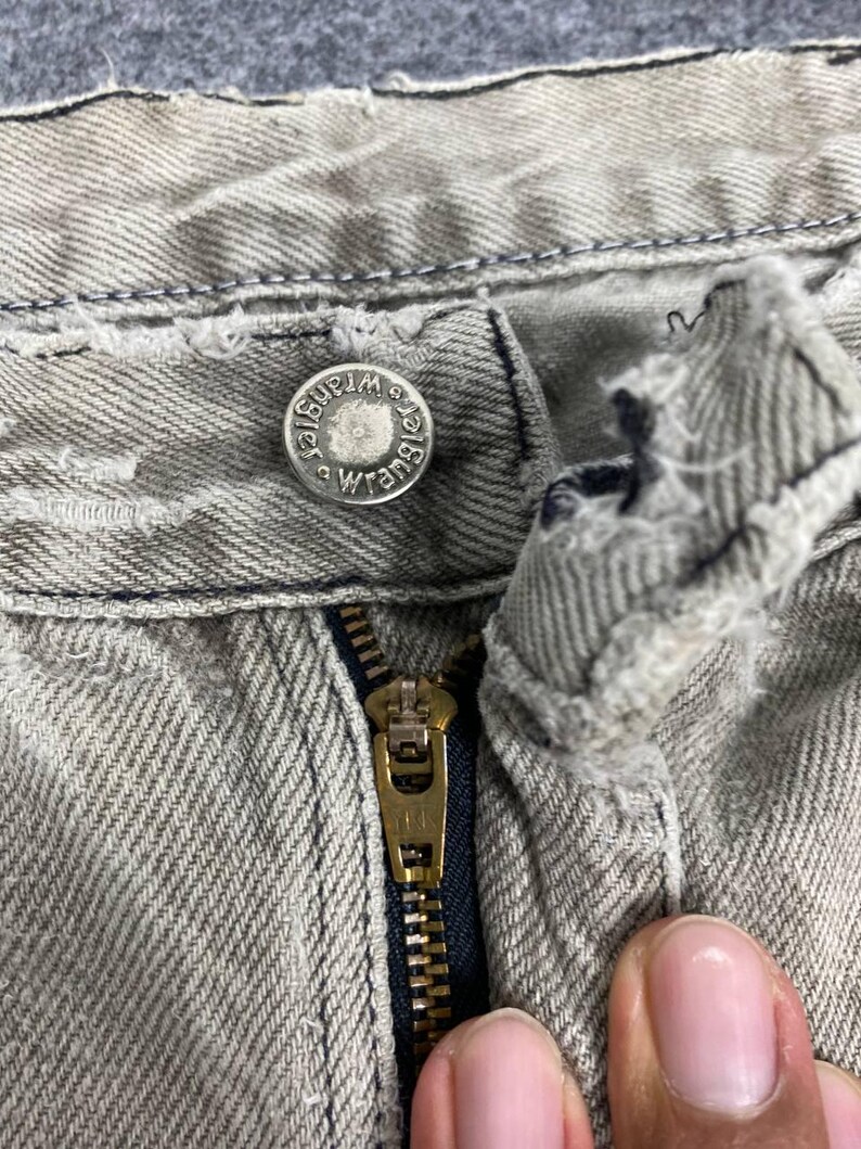 May include: Close-up of the top portion of a pair of light gray denim jeans. The jeans feature a metal button with the "Wrangler" logo, a brass zipper, and visible stitching details. The fabric shows signs of wear.
