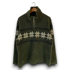 May include: An olive green fleece pullover with a high collar and a quarter-zip closure. The garment features a horizontal band across the chest decorated with a repeating cream and black snowflake pattern, set against the textured fleece fabric.