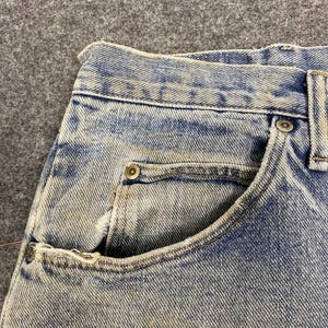 K&ouml;nnte beinhalten: Nahaufnahme einer hellblauen Jeans, die die Vordertasche und den Bund zeigt. Die Jeans hat ein verwaschenes, abgenutztes Aussehen mit sichtbaren N&auml;hten und Metallnieten. Der Stoff hat ein strukturiertes, leicht verwittertes Aussehen.