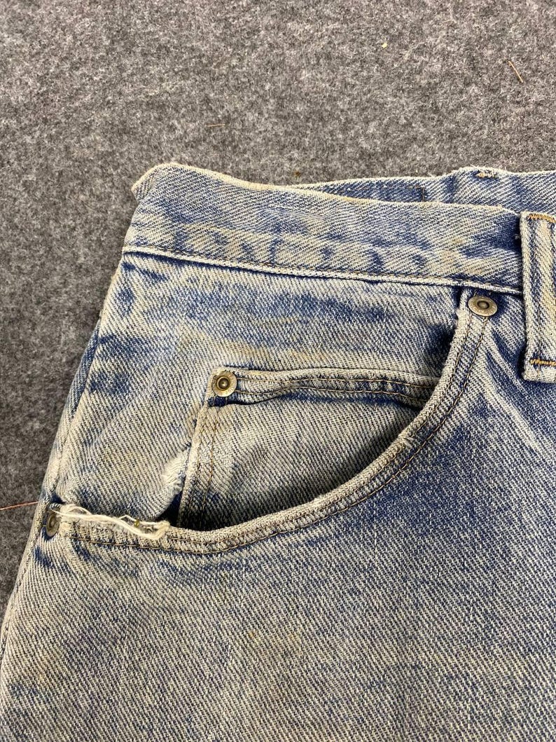 K&ouml;nnte beinhalten: Nahaufnahme einer hellblauen Jeans, die die Vordertasche und den Bund zeigt. Die Jeans hat ein verwaschenes, abgenutztes Aussehen mit sichtbaren N&auml;hten und Metallnieten. Der Stoff hat ein strukturiertes, leicht verwittertes Aussehen.