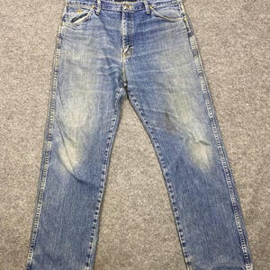 May include: Faded blue denim jeans with a classic five-pocket design. The jeans show signs of wear and fading, with a straight leg cut. The stitching is visible along the seams and pockets. The jeans are laid flat on a gray surface.